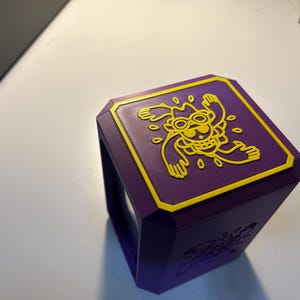 May include: A purple, geometric box with a yellow border and a yellow cartoon character design on the top. The box has a unique shape and appears to be 3D-printed. The design is a playful, whimsical illustration.