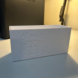 May include: A white rectangular storage box featuring a raised skull and crossbones design wearing a straw hat. The box is made of a solid white material and is on a white surface.