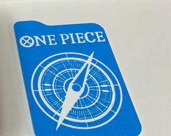 May include: A blue rectangular card with the words "ONE PIECE" in white, above a white compass design. The compass has a needle pointing upwards, and is surrounded by concentric circles. The card has a small notch at the top.