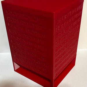 May include: A vibrant red, rectangular box with a lid. The box features embossed, ancient-looking text on all sides. The bottom section has a cutout. The box is made of a solid material and has a matte finish.