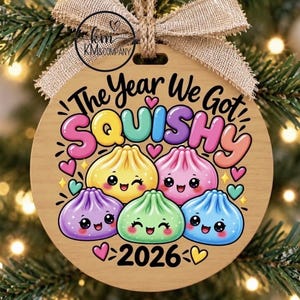 Puede incluir: Un adorno redondo de madera con un lazo de arpillera que presenta el texto The Year We Got SQUISHY y 2026. El diseño incluye cinco personajes de albóndigas coloridos y sonrientes en amarillo, rosa, morado, verde y azul, rodeados de corazones.