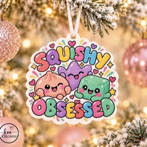 May include: A white scalloped ornament featuring a colorful cartoon illustration of a pink dumpling, a purple star, and a green cube with smiling faces. The text SQUISHY OBSESSED is printed in bold, multi-colored bubble letters around the characters.