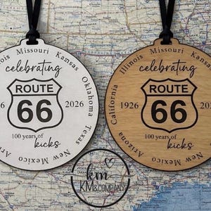 Peut inclure: Deux ornements circulaires Route 66. L'un est blanc, l'autre est en bois clair. Ils pr&eacute;sentent l'&eacute;cusson Route 66, les ann&eacute;es 1926 et 2026, et l'inscription "100 years of kicks". Suspendus par des cordons noirs.