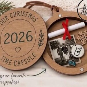 以下が含まれることがあります： 「OUR CHRISTMAS 2026 TIME CAPSULE」と刻印されたヒンジ付きの蓋を持つ、円形の木製クリスマス・タイムカプセル・オーナメント。内部には、赤いリボンで結ばれた白い紙の巻物、正方形の写真、木製の雪の結晶、小さな金属製の飛行機、青いスーツケースのチャーム、地図の輪郭とハートが描かれた円形の金属製チャームが入っています。