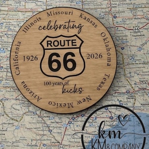 May include: A round wooden Route 66 sign with the text "celebrating Route 66" and "100 years of kicks." The sign has the years 1926 and 2026, and the names of states. The sign is on a map.