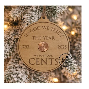 May include: A round wooden Christmas ornament with the text "IN GOD WE TRUST" and "WE LOST OUR CENTS". It features a penny and the years 1793 and 2025. The ornament hangs from a string and is set against a snowy tree.