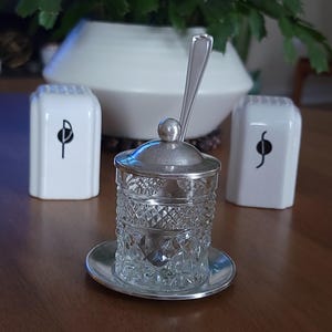 May include: A vintage glass and silver condiment set. The set includes a small, diamond-patterned glass jar with a silver lid and spoon, resting on a silver saucer. Two white salt shakers with black musical note designs are on either side.