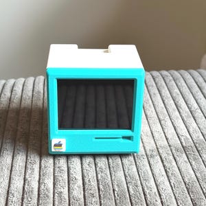 May include: A retro-style teal and white computer-shaped box with a black screen. The box has a slot at the bottom and a small logo on the front. The design is reminiscent of vintage computer aesthetics.