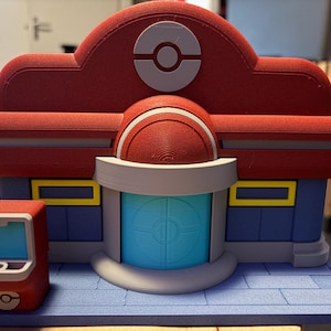 Op de afbeelding: Een 3D-geprint model van een Pokémon Center, met een rood dak met een wit Poké Ball-embleem. Het gebouw heeft blauwe muren, gele raamaccenten en een lichtblauwe deur. Links staat een kleine rode kiosk met een scherm.