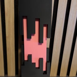 Könnte beinhalten: Ein hohes, rechteckiges Dekorationsobjekt in Schwarz und Rosa. Der schwarze Rahmen hat ein pixeliges Design mit ausgeschnittenen Formen, die ein rosafarbenes Innere freilegen. Der Sockel ist hellgrau. Das Objekt steht aufrecht.