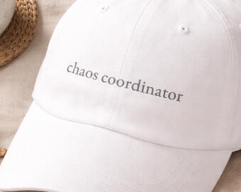 Puede incluir: Una gorra de béisbol blanca con el texto "chaos coordinator" en gris. La gorra está sobre una superficie beige, junto a una taza de café, gafas de sol y un libro.