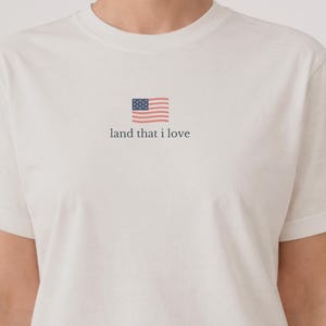 May include: White t-shirt with a small American flag graphic and the text "land that I love" in a simple font. The shirt has short sleeves and a crew neck.