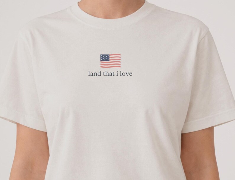 May include: White t-shirt with a small American flag graphic and the text "land that I love" in a simple font. The shirt has short sleeves and a crew neck.
