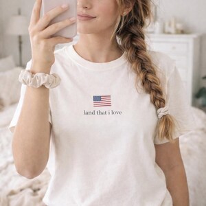 May include: White t-shirt featuring a small American flag graphic and the text "land that I love". The shirt is made of a soft material and has short sleeves. A scrunchie is visible on the wrist.