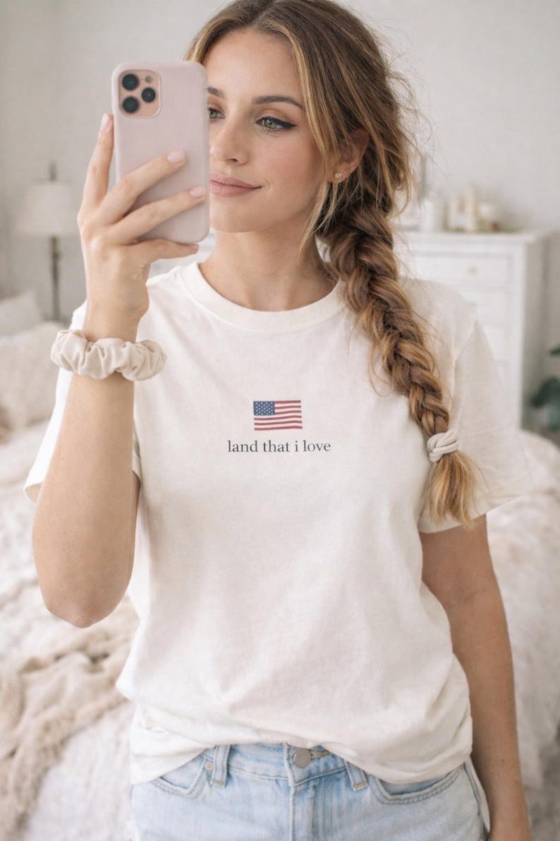 May include: White t-shirt featuring a small American flag graphic and the text "land that I love". The shirt is made of a soft material and has short sleeves. A scrunchie is visible on the wrist.