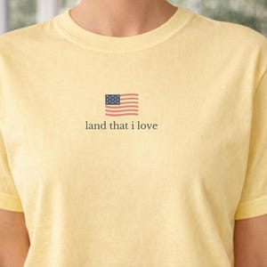 May include: A light yellow t-shirt with a small American flag graphic and the text "land that I love." The shirt is made of a soft material and has short sleeves. The design is centered on the chest.
