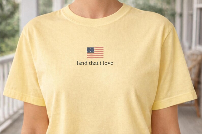 May include: A light yellow t-shirt with a small American flag graphic and the text "land that I love." The shirt is made of a soft material and has short sleeves. The design is centered on the chest.