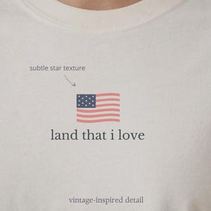 May include: Cream-colored t-shirt featuring a small American flag graphic with the text "land that I love." The flag is red, white, and blue, with a subtle star texture and vintage-inspired detail.