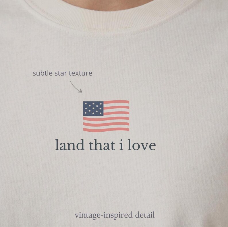 May include: Cream-colored t-shirt featuring a small American flag graphic with the text "land that I love." The flag is red, white, and blue, with a subtle star texture and vintage-inspired detail.