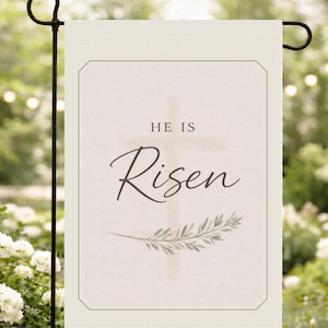 May include: A cream-coloured garden flag featuring the text "HE IS Risen" in cursive, a cross, and a sprig of leaves. The flag has a thin border and is displayed on a black metal stand, set against a blurred outdoor backdrop.