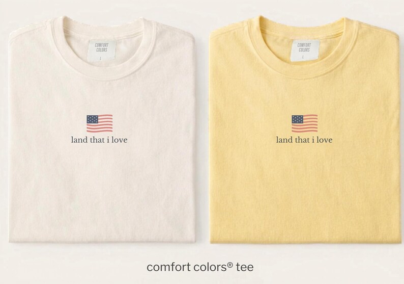 May include: Two Comfort Colors tees, one cream and one yellow, each with a small American flag graphic and the text "land that I love." The shirts are folded and displayed side-by-side, showcasing a patriotic theme.