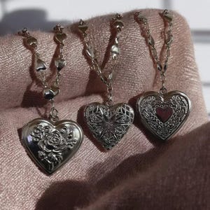 May include: Three silver heart-shaped lockets with intricate designs. One locket features a rose, another a starburst, and the third a floral pattern with a red center. Each locket hangs from a delicate chain with heart-shaped accents.