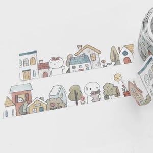 May include: A roll of decorative washi tape with a repeating pattern of cartoon houses, trees, and characters. The tape is partially unrolled, showing the design. A pink and white striped pen and a blue pen are also visible.