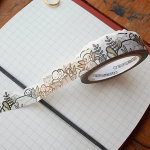 May include: Washi tape with a forest-themed design in brown, green, and beige. The tape roll displays a repeating pattern of cartoon characters, trees, and mushrooms. The text "THECOFFEEMONSTERCO" is printed on the tape. The tape is partially unrolled.