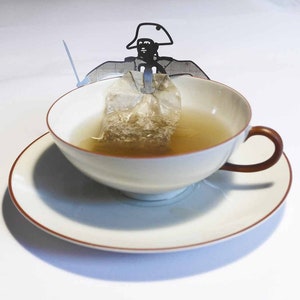 Könnte beinhalten: Eine weiße Teetasse und Untertasse mit braunem Rand. Ein Teebeutel ist im Tee versunken, und ein Teebeutelhalter aus Metall in Form einer Person mit Hut befindet sich oben auf der Tasse.