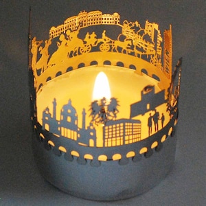 May include: A silver candle holder with a cut-out design of a city skyline. The candle holder is lit with a candle inside, casting a warm glow.