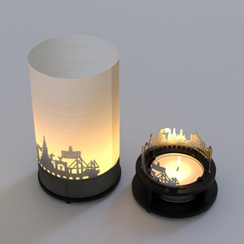 May include: Two candle holders with a cityscape silhouette design. The larger holder is a white cylinder with a black base. The smaller holder is a black circular base with a cityscape silhouette around the top. A lit tea light candle is in the smaller holder.