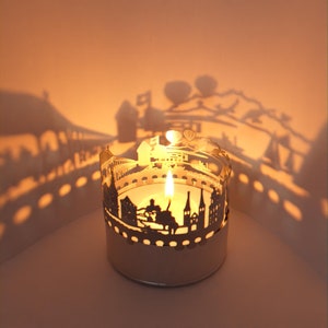 Puede incluir: Un portavelas de metal blanco con un diseño recortado de un horizonte de ciudad. El portavelas está iluminado con una vela en su interior, proyectando un cálido resplandor.