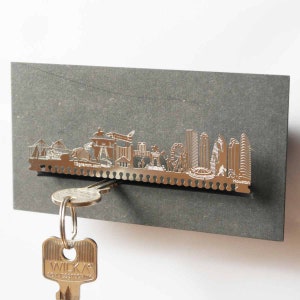 May include: A silver metal key holder shaped like the Houston skyline. The key holder is mounted on a gray background. A silver key is hanging from the holder.