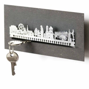 Può includere: Un portachiavi in metallo argentato a forma di skyline di Norimberga. Il portachiavi ha una fessura per le chiavi e la parola "Norimberga" è incisa in basso. Una chiave è appesa al portachiavi.