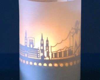 May include: A white paper candle holder with a cityscape silhouette design. The silhouette is cut out of the paper, allowing light to shine through.