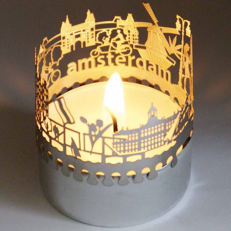 May include: A silver candle holder with a cut-out design of the Amsterdam skyline. The candle holder features a lit tea light candle inside. The text "amsterdam" is visible on the candle holder.