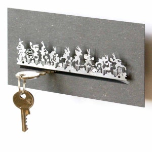 May include: Silver key holder with a design of a line of rabbits holding Easter eggs. The key holder is mounted on a gray background. A silver key is hanging from the holder.