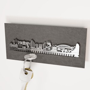 May include: A silver metal key holder shaped like the city skyline. The key holder is mounted on a dark grey wall. A key is hanging from the key holder.