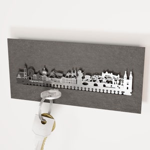 May include: A silver metal key holder with a city skyline silhouette, attached to a dark grey rectangular board. The key holder has small hooks along the bottom. The website address 13gramm.com is visible. A set of keys hangs from the key holder.