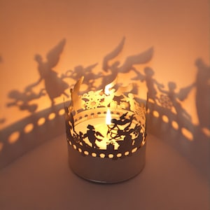 May include: A white metal candle holder with a cutout design of angels and a snowflake. The candle holder is lit and casting shadows of the angels on the wall.