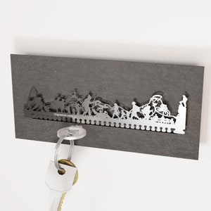 May include: A silver metal key holder with a silhouette of a group of people walking. The key holder is mounted on a dark gray wall. A gold key is hanging from the key holder.