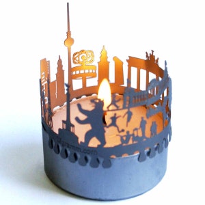 K&ouml;nnte beinhalten: Ein schwarzer Kerzenhalter aus Metall in Form der Berliner Skyline. Der Halter zeigt ber&uuml;hmte Sehensw&uuml;rdigkeiten wie das Brandenburger Tor, die Berliner Mauer und den Berliner B&auml;ren. Eine brennende Kerze befindet sich im Halter.