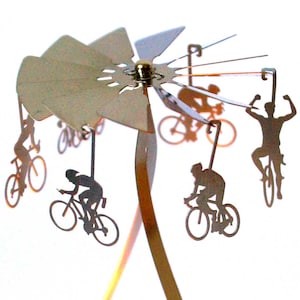 May include: A silver metal mobile with five silhouettes of cyclists riding bicycles. The mobile is suspended from a silver metal base.