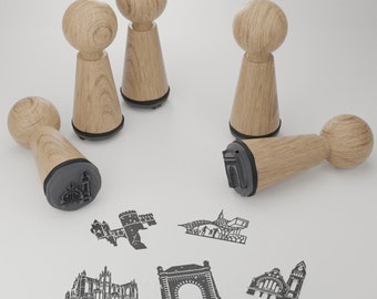 May include: Five wooden stamps with black ink pads. The stamps are shaped like a cylinder with a rounded top. The stamps have different designs, including an archway, a building, and a stylized design.