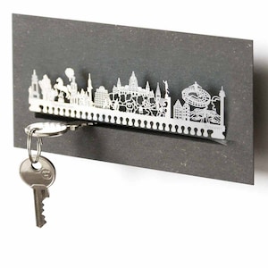 May include: A silver metal key holder shaped like the Hannover skyline. The holder is mounted on a gray wall and has a key attached to it.