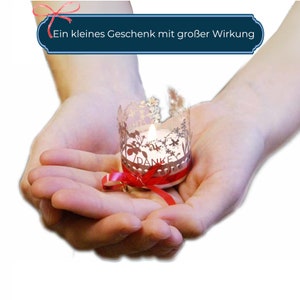 May include: A small, white candle with a delicate, laser-cut design featuring flowers and the word "Danke" is held in cupped hands. The candle is tied with a red ribbon.