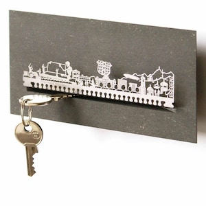 May include: A silver metal key holder shaped like the city skyline of Bregenz, Austria. The key holder is mounted on a gray background and has a key attached to it.