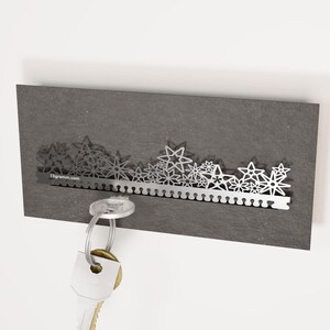 May include: A silver key holder with a decorative design of stars and flowers. The key holder is mounted on a dark gray wall. The text "13gramm.com" is visible on the key holder.