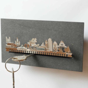 Puede incluir: Un porta llaves de metal plateado con forma de horizonte de Montreal. El soporte está montado sobre un fondo de pizarra gris. Una llave cuelga del soporte.