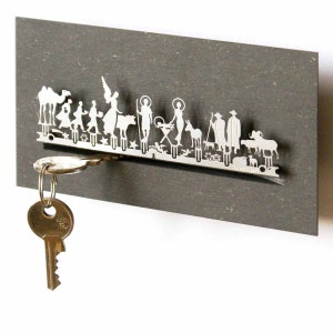 May include: A silver metal key holder shaped like a nativity scene with figures of people, animals, and a star. The key holder is mounted on a gray background and has a key attached to it.